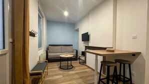 Un espacio habitable compacto con un sofá, una pequeña mesa redonda, un televisor y una zona de bar con taburetes.