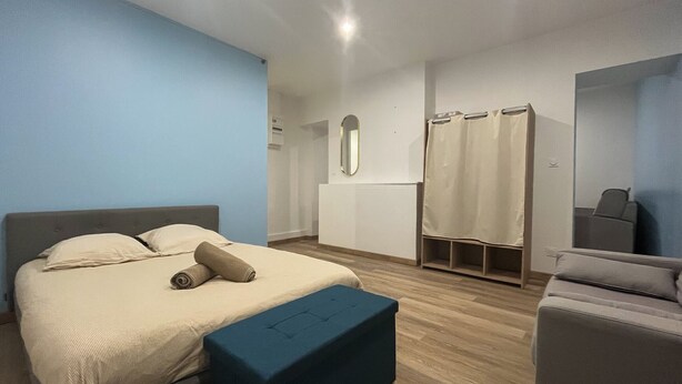 Un dormitorio con una cama, un banco azul, un sillón gris, un espejo y una zona de ducha.