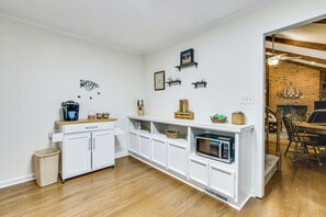 Eine Küche mit weißen Schränken, einer Mikrowelle und einer Kaffeemaschine. Es gibt Regale mit Dekorationen sowie einen Mülleimer.