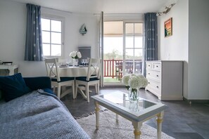Ein Zimmer mit einem Bett, einem Esstisch mit Stühlen, einem kleinen Tisch mit einer Vase, einer Kommode und einer Schiebetür zu einem Außenbereich.