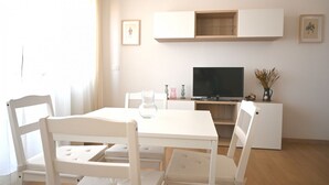 Ein weißer Esstisch mit vier Stühlen, ein Flachbildfernseher, eine weiße Kommode und eine Vase mit Trockenpflanzen.