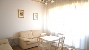 Ein Zimmer mit einem weißen Sofa, einem kleinen runden Tisch, einem weißen Esstisch mit vier Stühlen und zwei gerahmten Bildern an der Wand.
