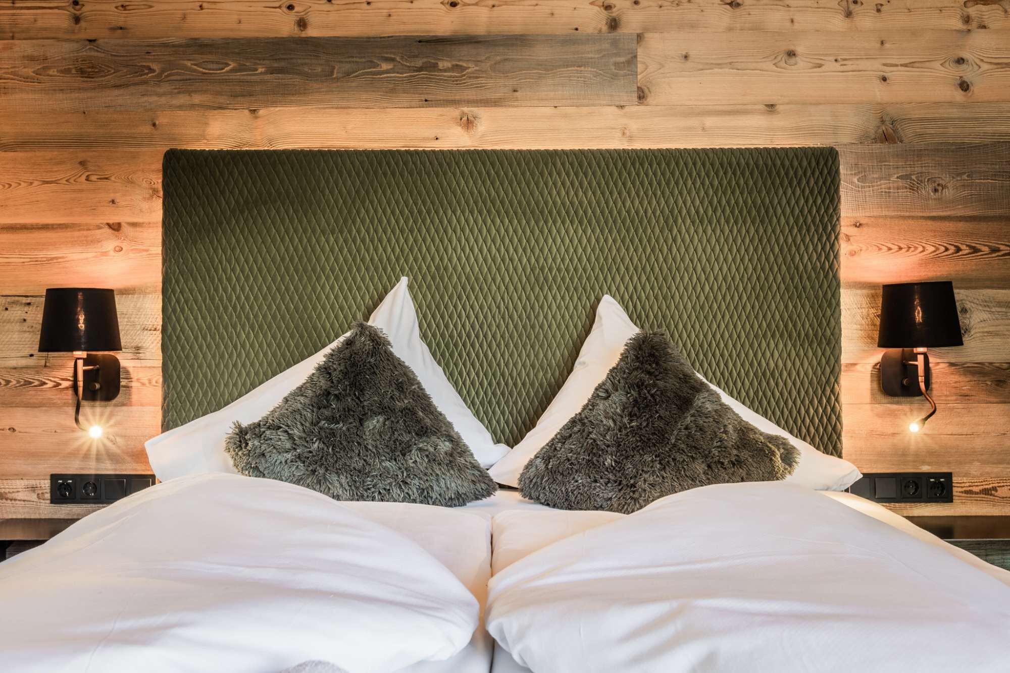 Una cama con cabecera acolchada verde, dos almohadas mullidas y dos lámparas fijadas a la pared.