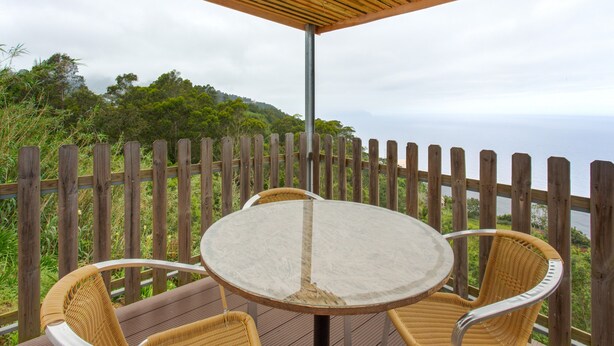 A wooden deck with a round table and wicker chairs, overlooking a forested hillside. - Sunset Village A Apartment with Sea View, Wi-Fi and Air Conditioning (Sao Jorge)