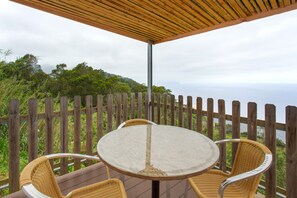 A wooden deck with a round table and wicker chairs, overlooking a forested hillside. - Sunset Village A Apartment with Sea View, Wi-Fi and Air Conditioning (Sao Jorge)