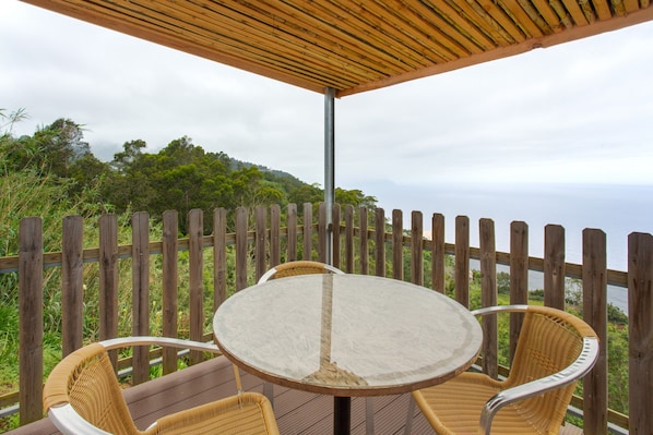 Eine Holzterrasse mit einem runden Tisch und Korbstühlen, mit Blick auf einen bewaldeten Hang.