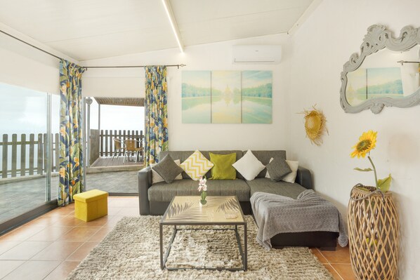 A living room with a sofa, a coffee table, a yellow ottoman, a large mirror, and a view of a balcony.