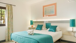 Ein ordentlich bezogenes Bett mit himmelblau gestreifter Tagesdecke und Kissen, eine Nachttischlampe, ein gerahmtes Bild an der Wand und ein Fenster mit Vorhängen.