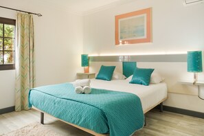Ein ordentlich bezogenes Bett mit himmelblau gestreifter Tagesdecke und Kissen, eine Nachttischlampe, ein gerahmtes Bild an der Wand und ein Fenster mit Vorhängen.