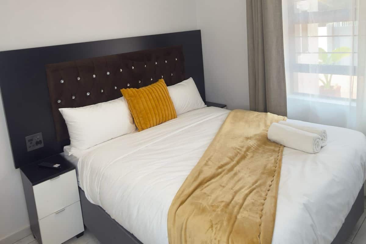 Una cama bien hecha con cabecera, una mesita de noche con cajón y una ventana con cortinas.