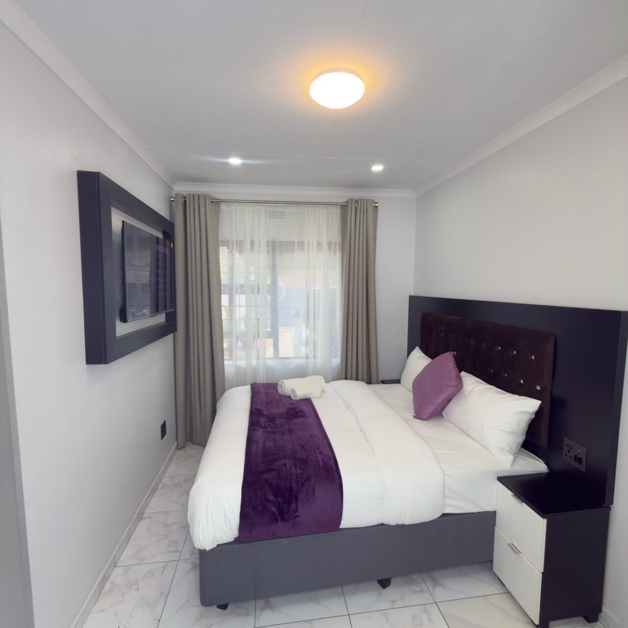A modern bedroom with a bed, a nightstand, a window with curtains, and a ceiling light.