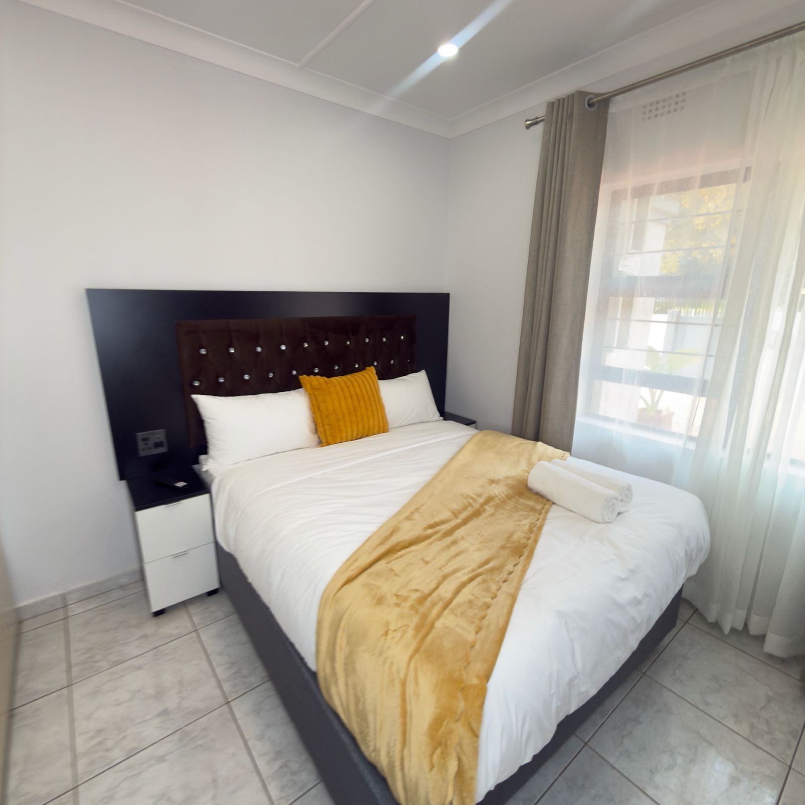 A hotel room with a bed, a nightstand, a drawer, a window with curtains, and a ceiling light.