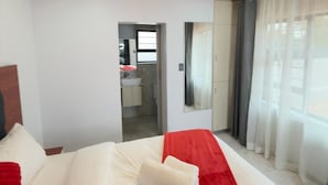 A bedroom with a wooden ceiling, a bed with white and red bedding, a bathroom visible through an open door, and a large window with curtains. - Cozy Villa Lodge (Edenvale)