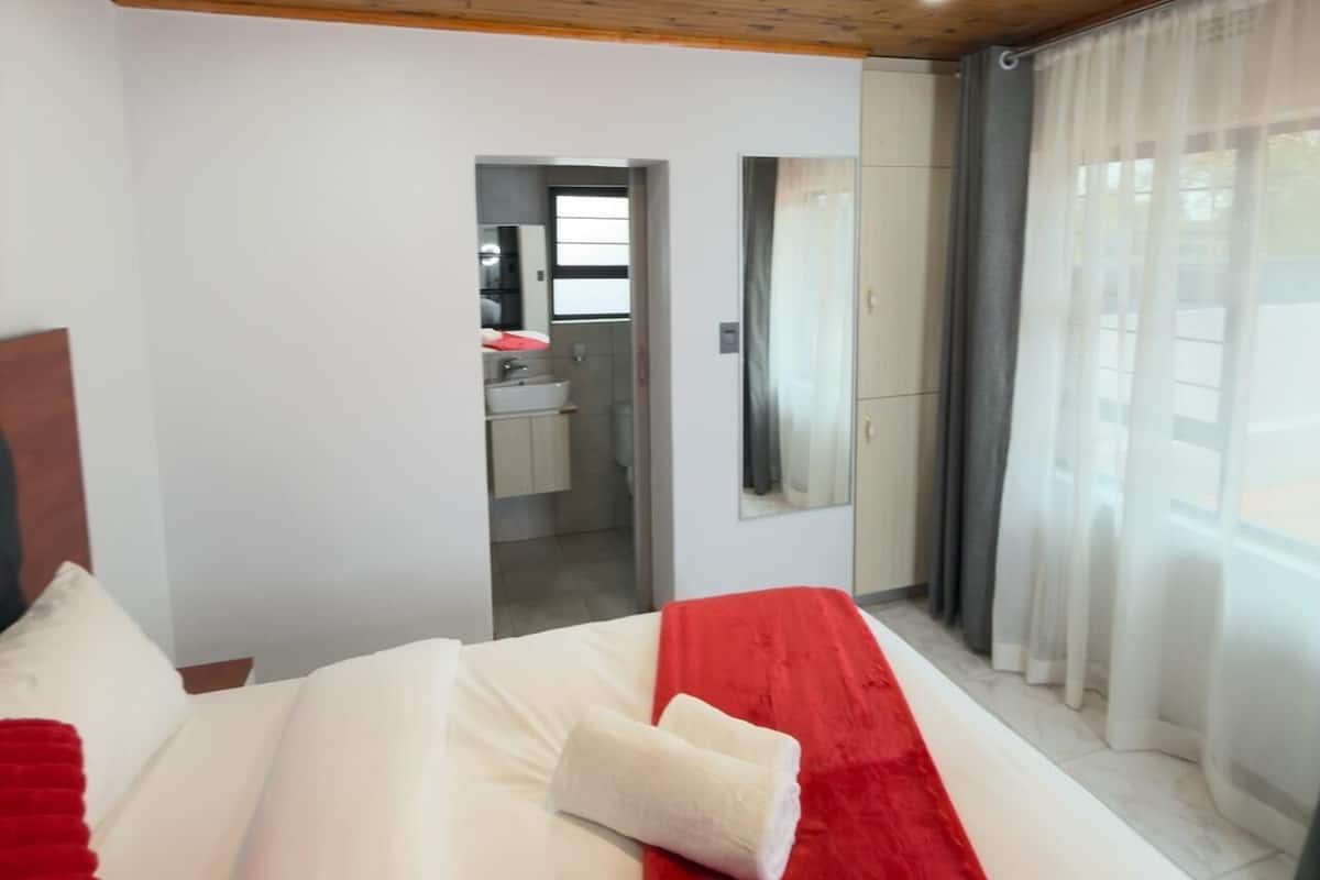 Un dormitorio con techo de madera, una cama con ropa de cama blanca y roja, un baño visible a través de una puerta abierta y una ventana grande con cortinas.