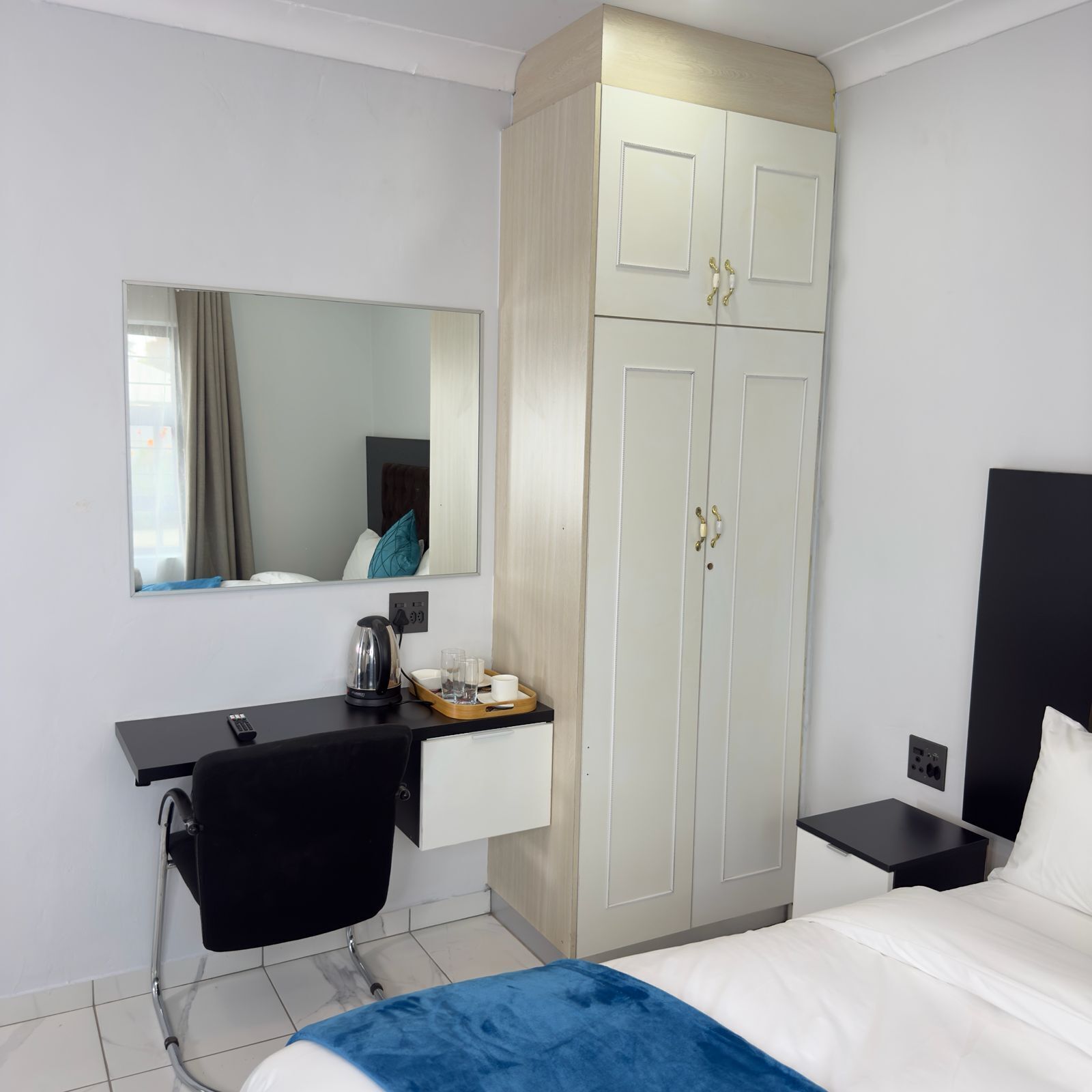 A hotel room with a bed, a desk with a chair, a wardrobe, a mirror, and a bedside table with a lamp.