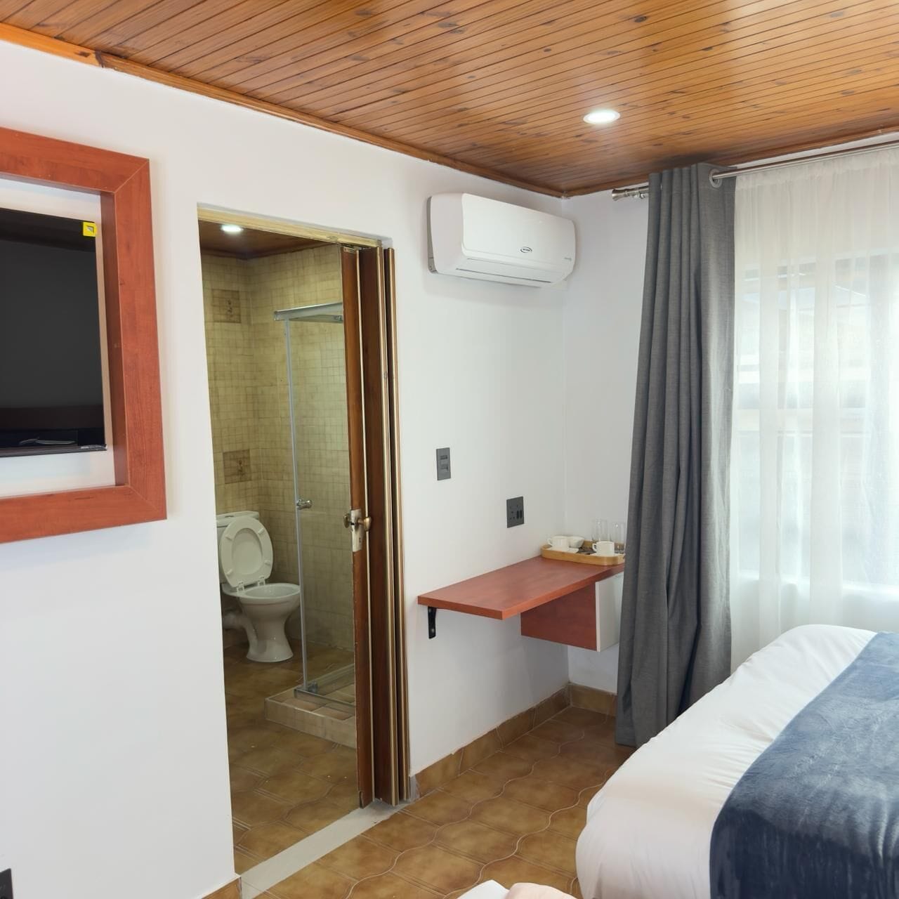 A hotel room with a wooden ceiling, a mounted air conditioner, a shower area, a toilet, a bed with a blue blanket, and a small wooden shelf.