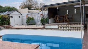 A swimming pool with a tiled deck, a covered patio area with chairs, and a small shed in the background. - Cozy Villa Lodge (Edenvale)