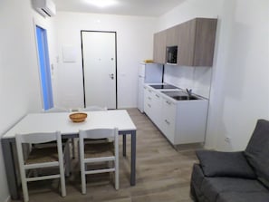 Eine kompakte Küche mit weißen Schränken, einer integrierten Mikrowelle und einem Spülbecken. Im Wohnbereich befindet sich ein Esstisch mit Stühlen und ein graues Sofa.