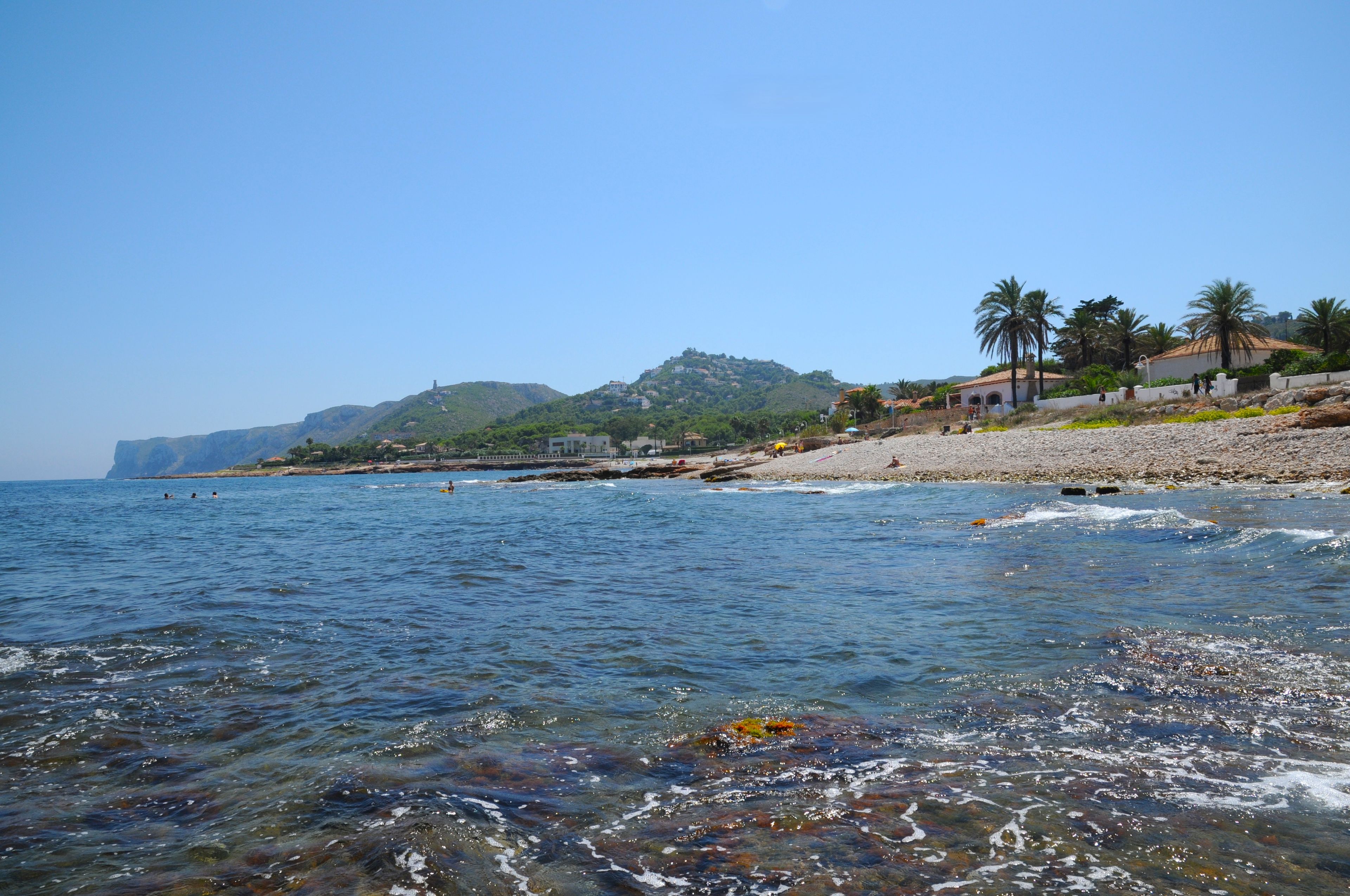 A coastal beach with clear water, rocky shore, and a hill with buildings in the background.