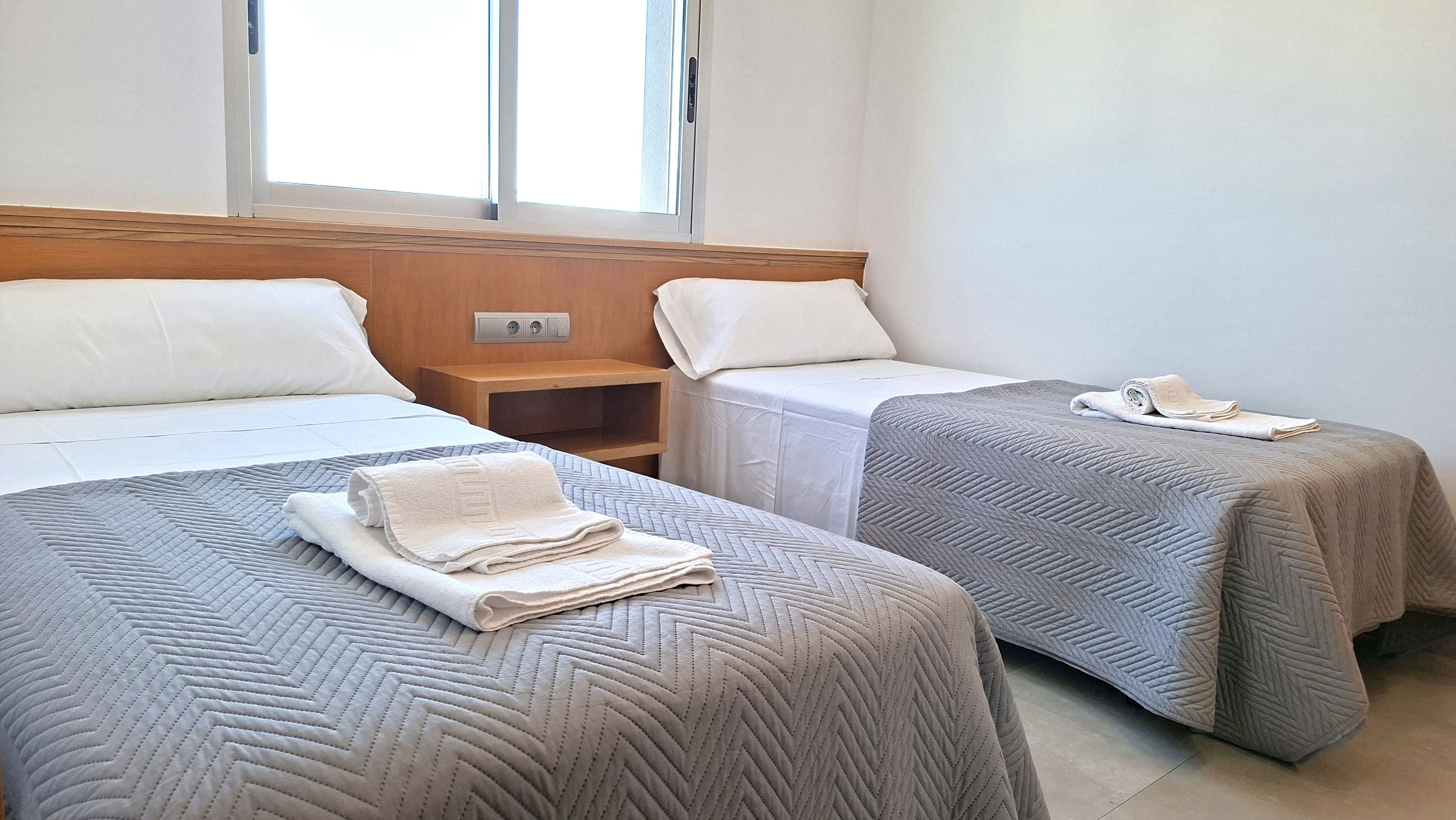 A hotel room with two beds, each with a pillow and a folded towel. There is a wooden headboard, a nightstand with a lamp, and a window with blinds.