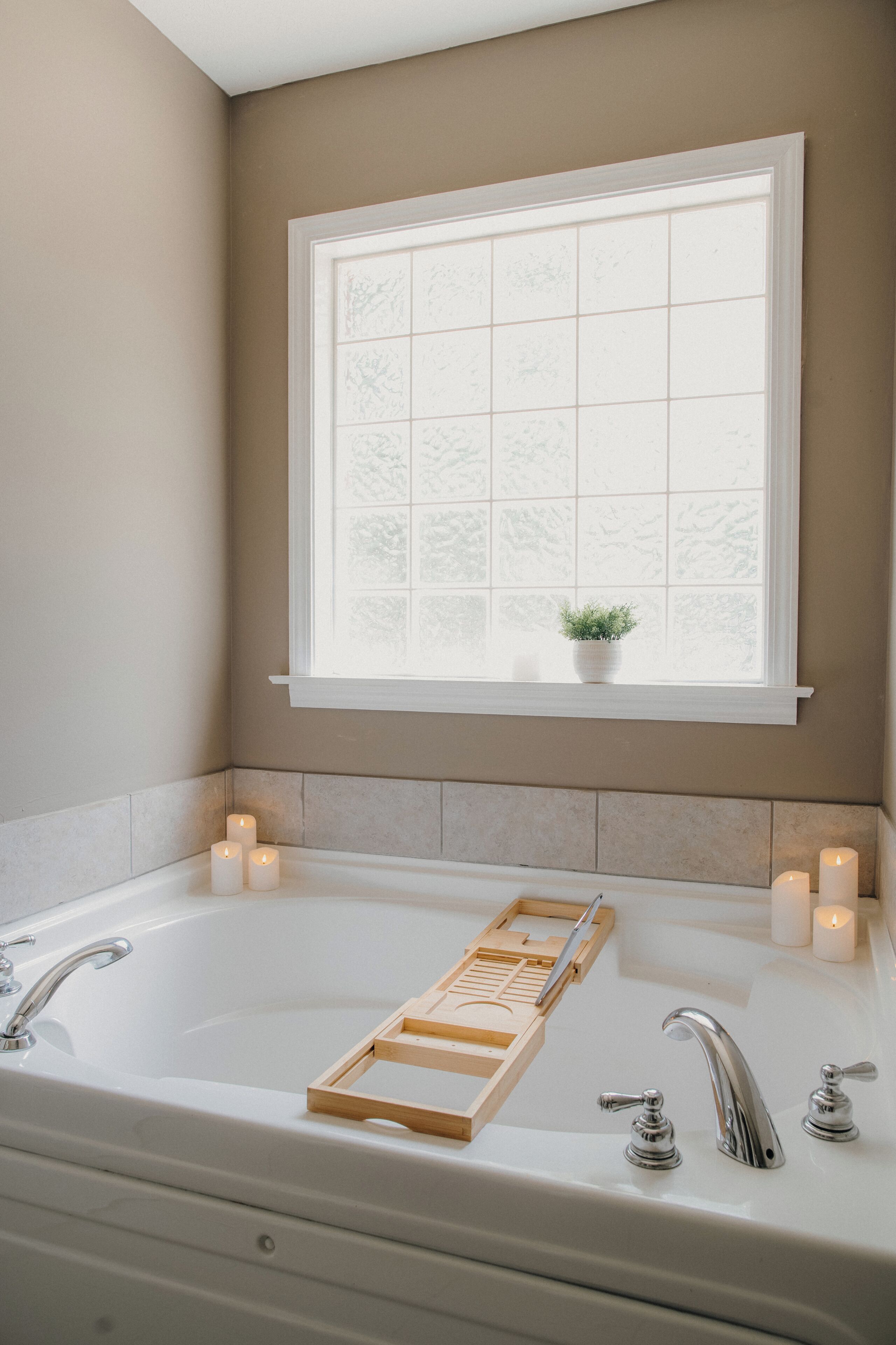 A bathroom with a bathtub, two faucets, a window with a plant, and three lit candles.
