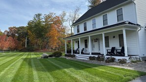 A white two-story house with a porch, a well-maintained lawn, and autumn-colored trees in the background.