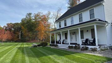 A white two-story house with a porch, a well-maintained lawn, and autumn-colored trees in the background.