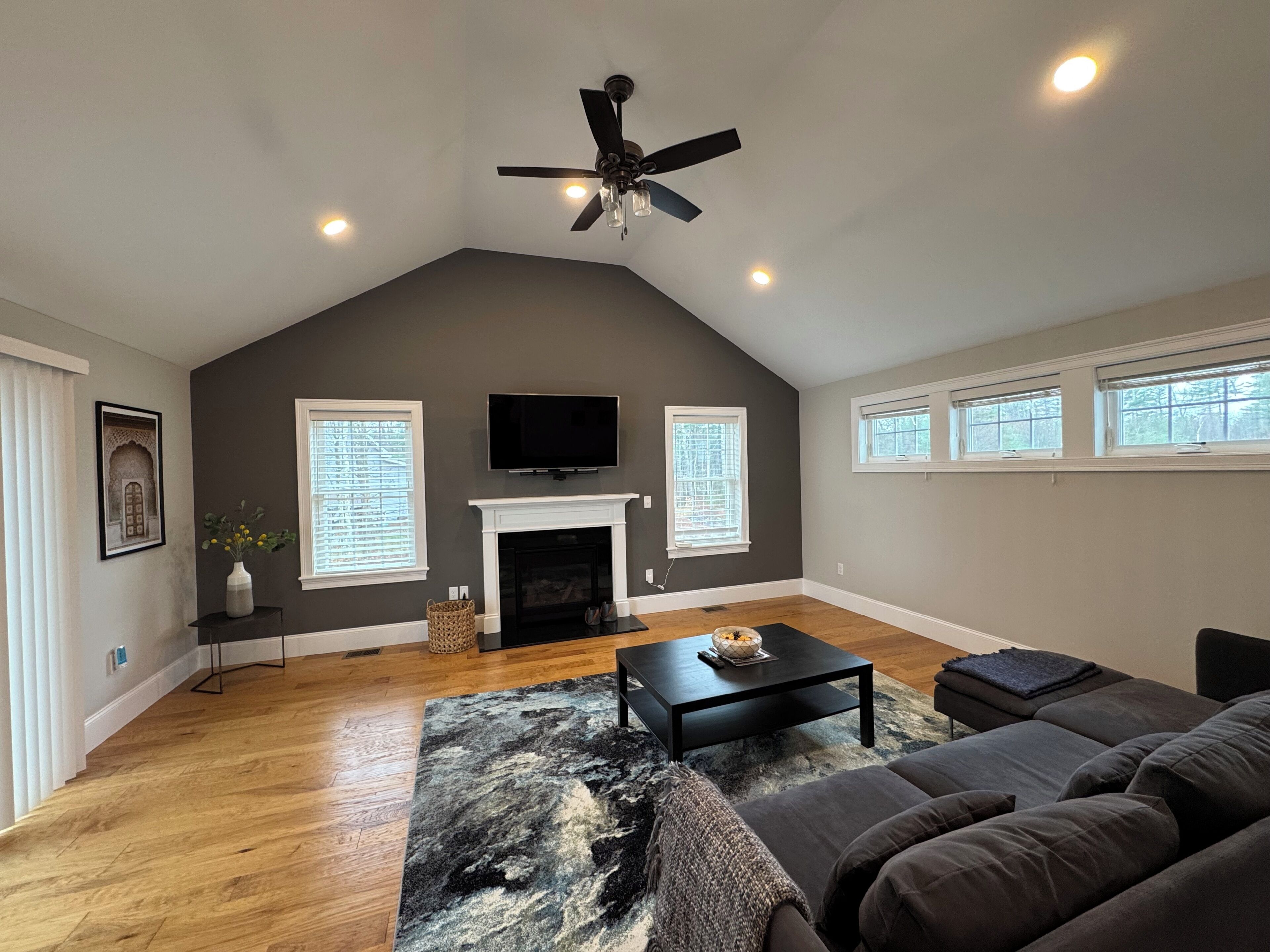 A spacious living room with a fireplace, a ceiling fan, and a large window.