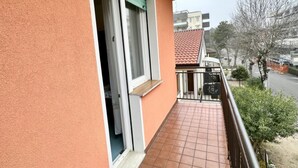 Un balcón con piso de baldosas, una ventana con cortina y vista a una calle con edificios y árboles.