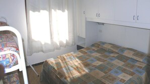 Pieni huone, jossa on yksi sänky, pelkistetty sängynpeite, valkoinen vaatekaappi ja ikkuna, jossa on ohuet verhot.