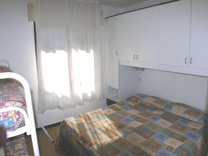 A small, single-bed room with a plain bedspread, a white wardrobe, and a window with sheer curtains. - Functional stay for five guests in Bibione (Bibione)