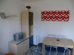 Une pièce avec une table en bois, quatre chaises, un téléviseur posé sur un meuble et un élément de décoration murale rouge à motifs.