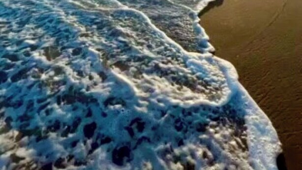 Uma praia com ondas, céu claro e o sol se pondo ou nascendo.