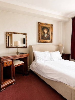 Quarto de hotel com uma cama, uma escrivaninha com luminária, uma cadeira, um espelho e um retrato emoldurado na parede.