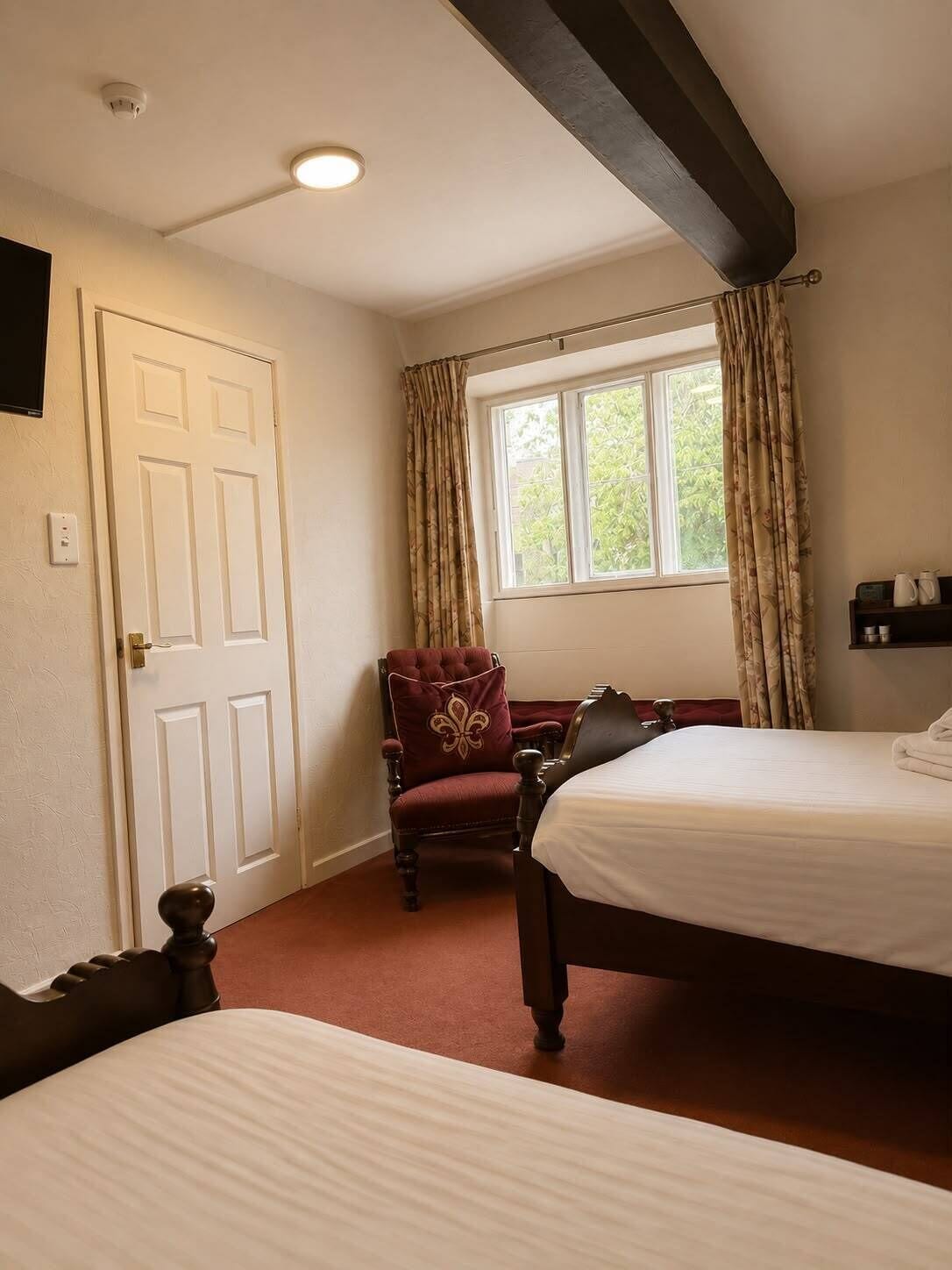 A hotel room with two beds, a chair, a window with curtains, a door, and a ceiling light.