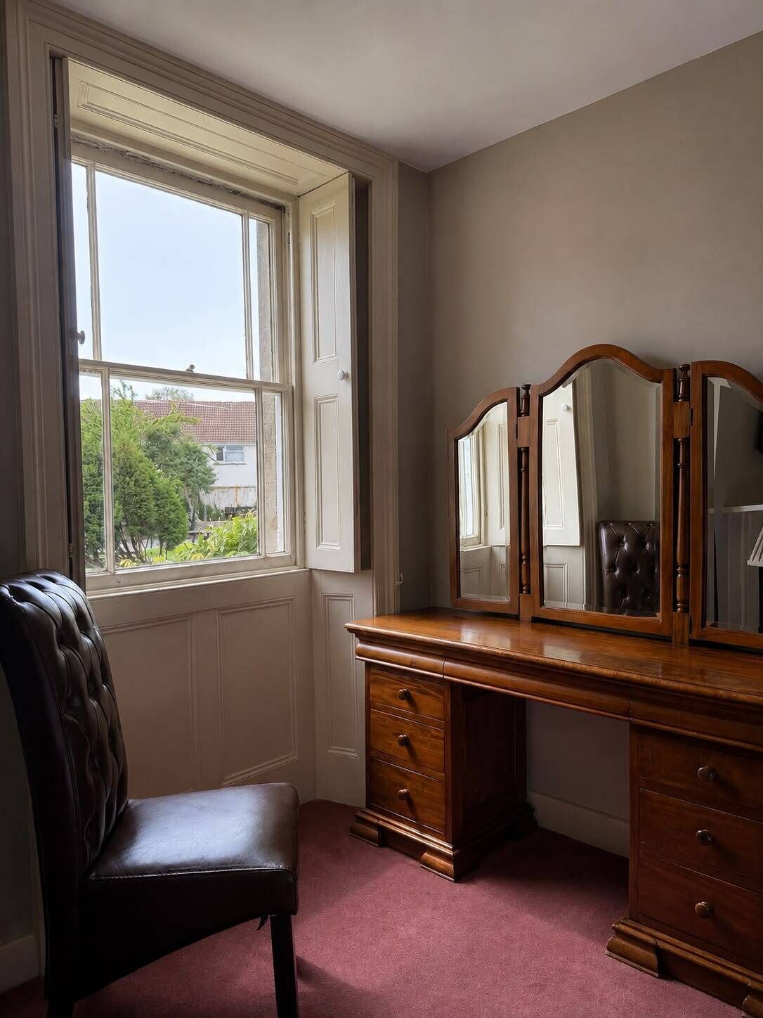 A room with a large window, a wooden dresser with drawers, a leather chair, and a mirror.