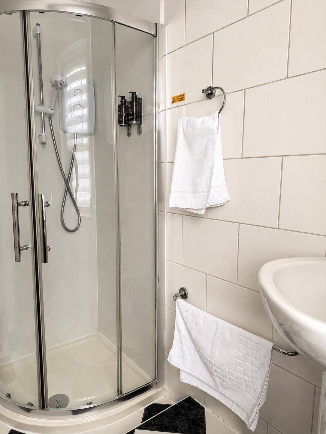 Une douche fermée par des parois de verre, dotée d’un pommeau de douche à main, d’un robinet mural et d’un sol carrelé.