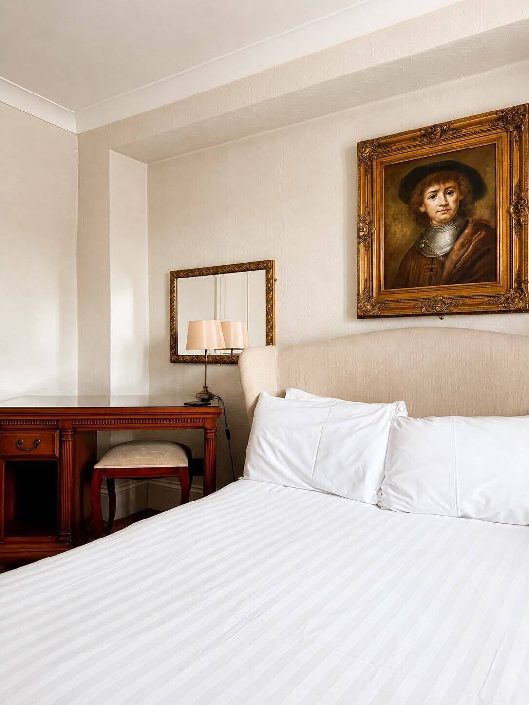 A hotel room with a bed, a desk, a chair, a lamp, and a framed portrait on the wall.