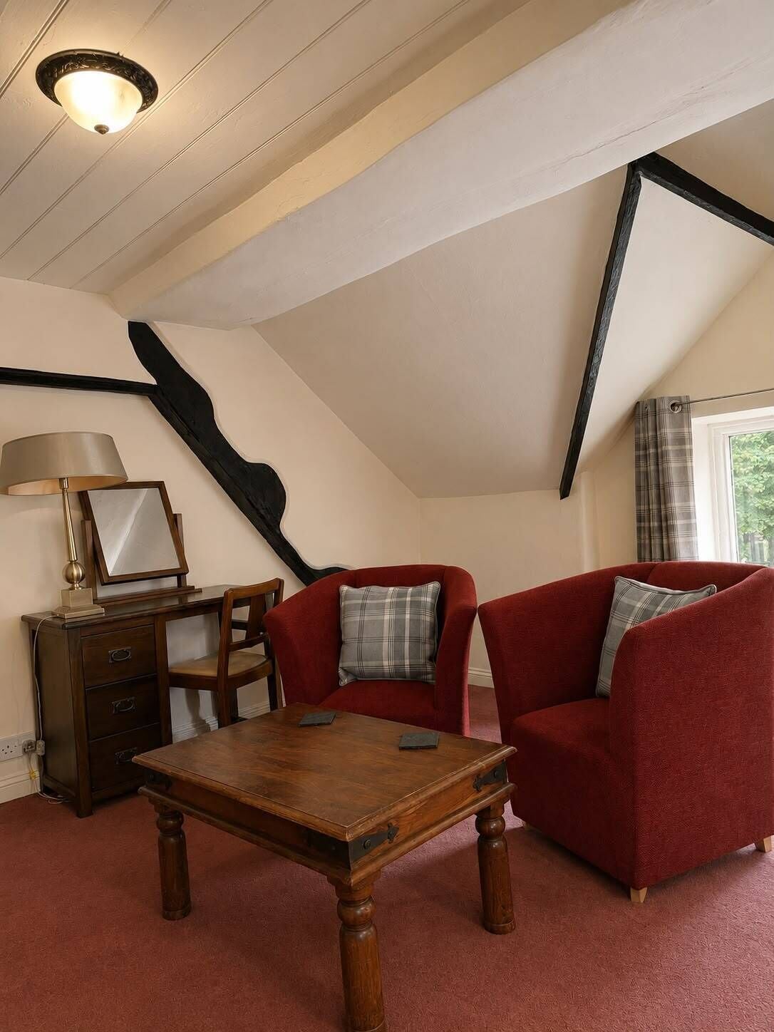A room with a sloped ceiling, a wooden table, two red armchairs, a small desk with a lamp, and a window with curtains.