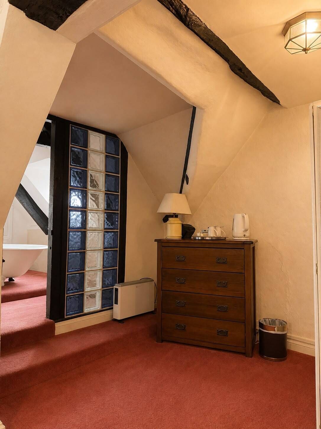 Una habitación con alfombra roja, cómoda de madera, lámpara, radiador y ventana con vidrios azules y blancos.