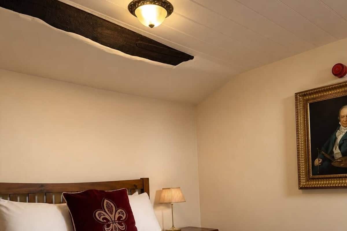 A hotel room with a large bed, a wooden headboard, a nightstand with a lamp, and a framed portrait on the wall.