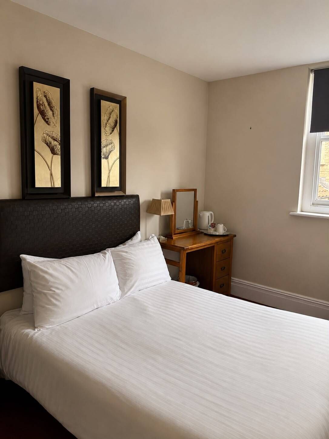Une chambre d’hôtel avec un lit, deux tableaux représentant des motifs floraux encadrés, une table de chevet en bois, une lampe et une fenêtre.
