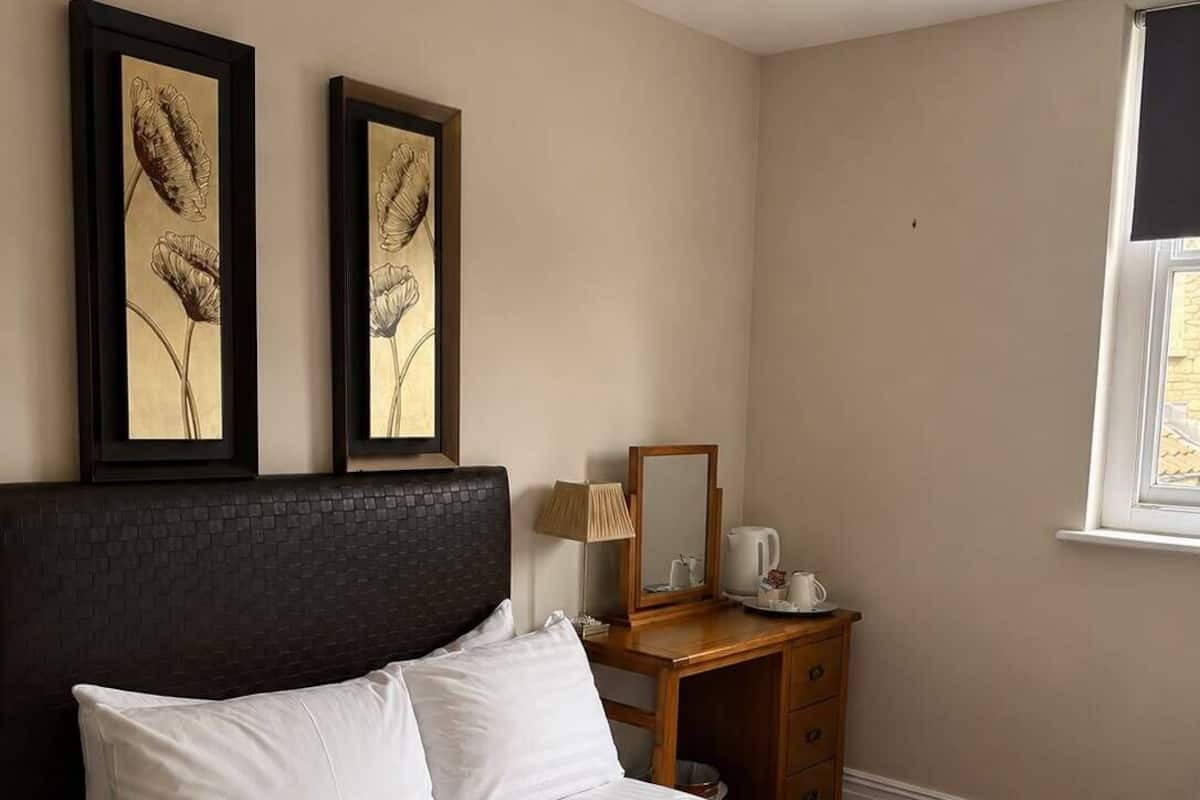 A hotel room with a bed, two framed floral paintings, a wooden nightstand, a lamp, and a window.