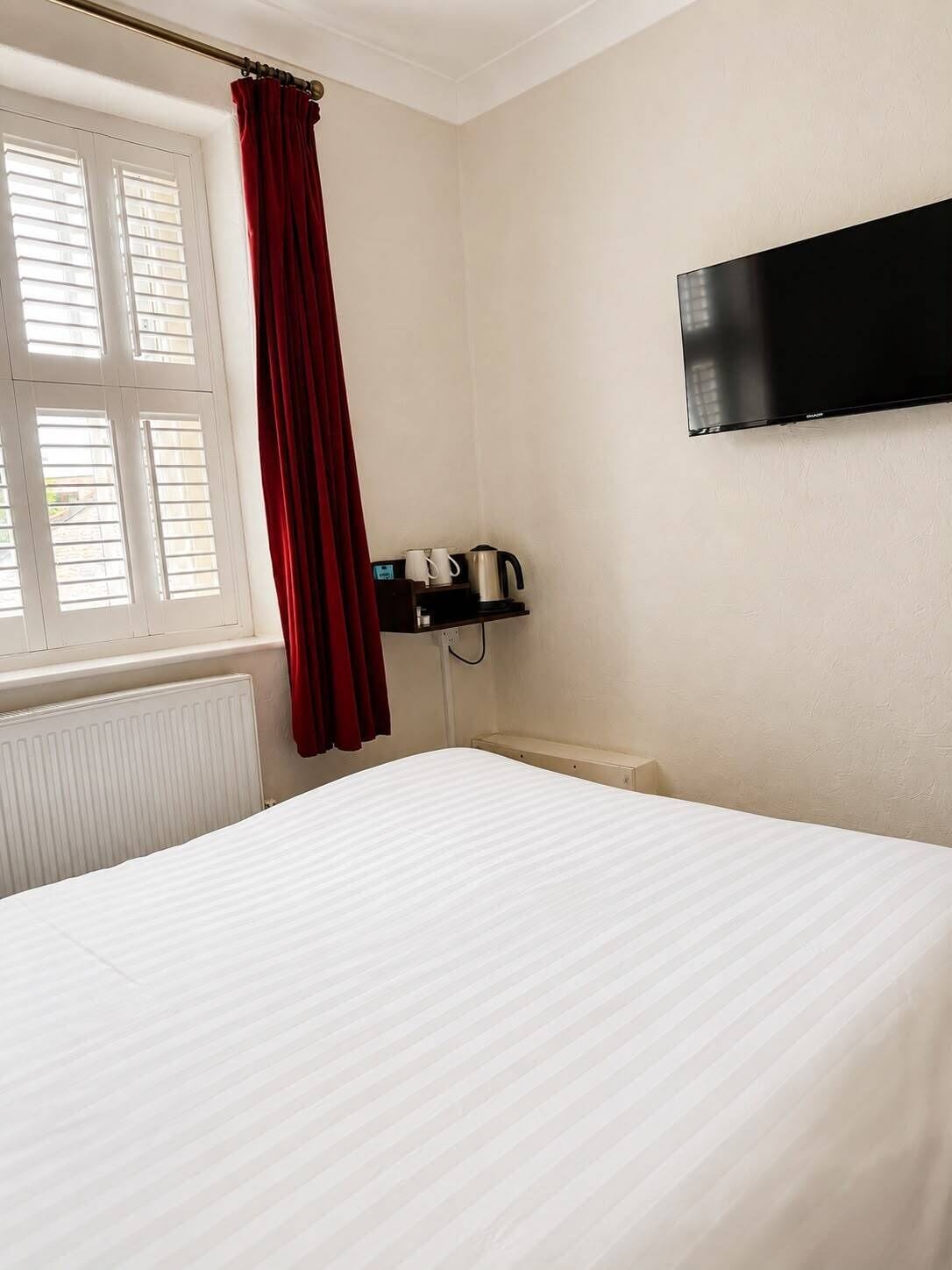 A hotel room with a bed, a TV mounted on the wall, a window with blinds, and a small shelf with a kettle and cups.
