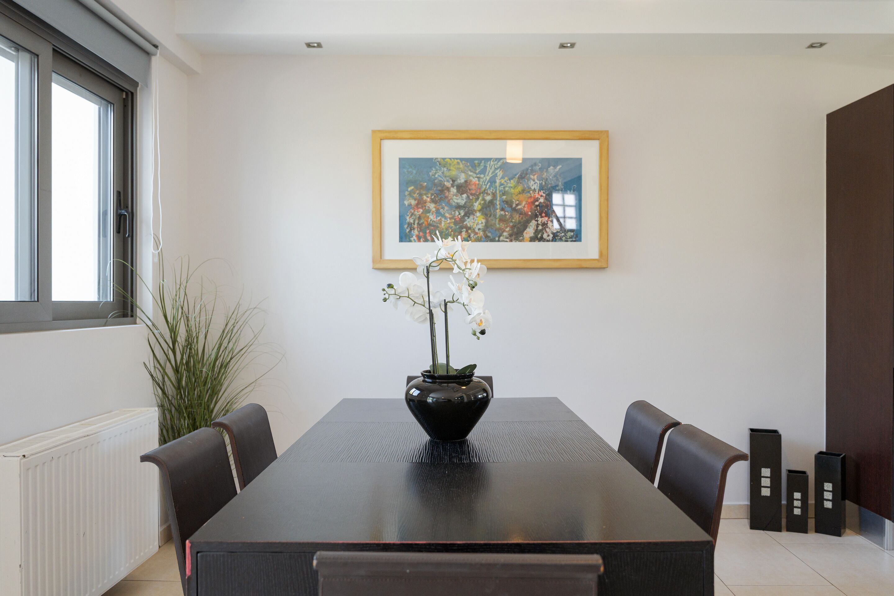 Un comedor con una mesa rectangular, sillas, un florero con flores, un cuadro en la pared y un radiador.