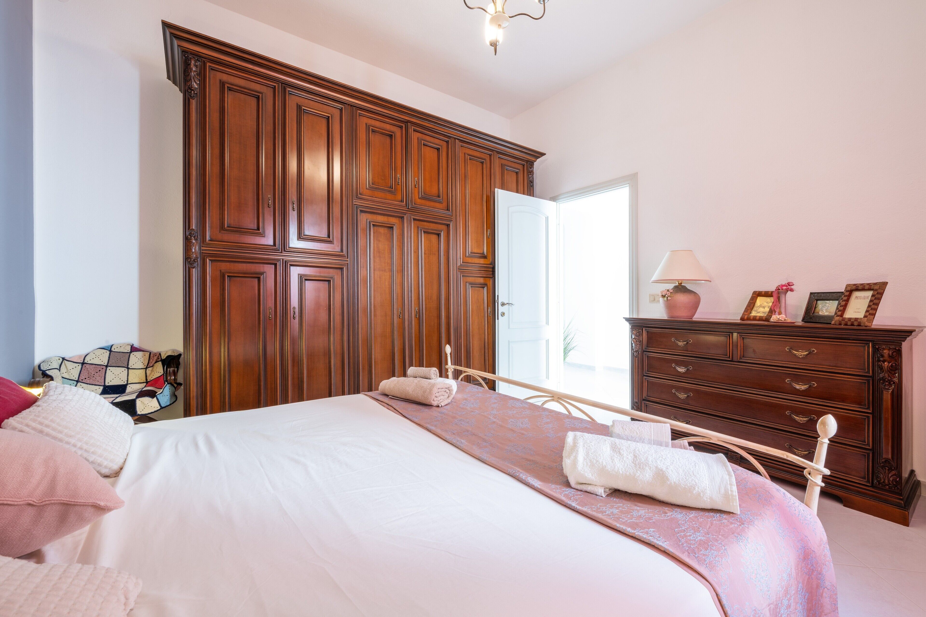 Un dormitorio con un armario grande de madera, una cama con colcha rosa, una mesita de noche con lámpara y un aparador con espejo.