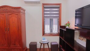 Quarto com guarda-roupa de madeira, televisão, uma pequena mesa com uma garrafa de água e janela com persianas.