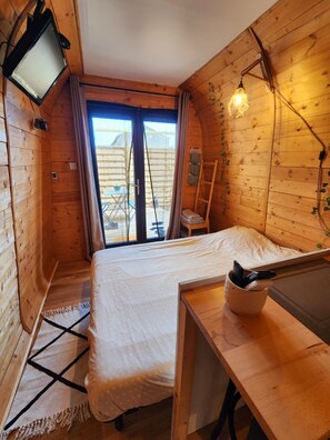 Ein gemütlicher Holzraum mit einem Bett, einem kleinen Tisch, einem Fernseher und Blick durch eine Glastür.