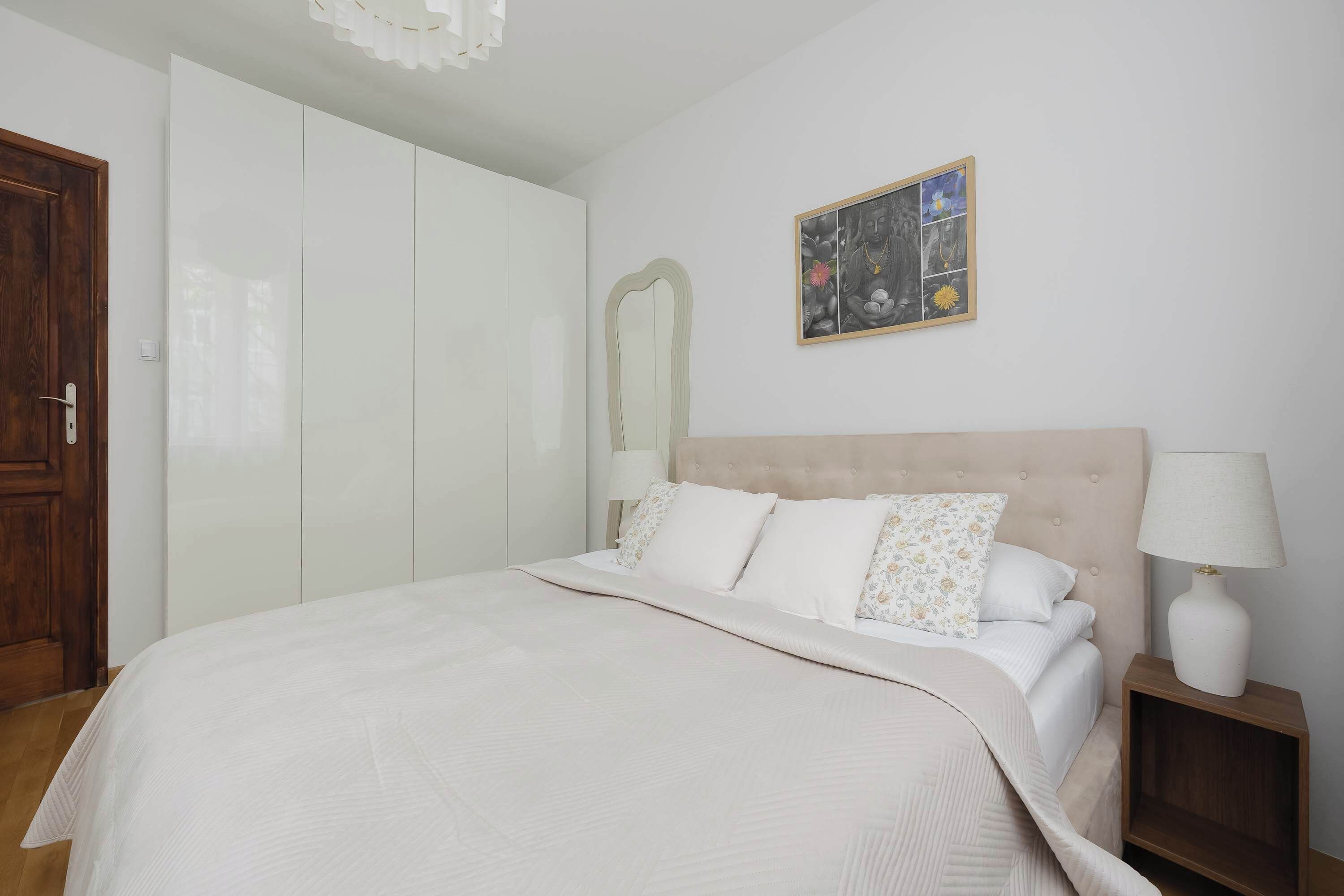 A bedroom with a bed, a nightstand, a lamp, a wardrobe, and a wall-mounted artwork.