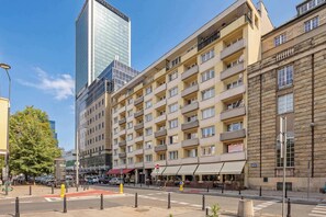 Ein mehrstöckiges Hotel mit einem modernen Glasgebäude im Hintergrund, eine Straße mit Autos und einer Fußgängerampel.